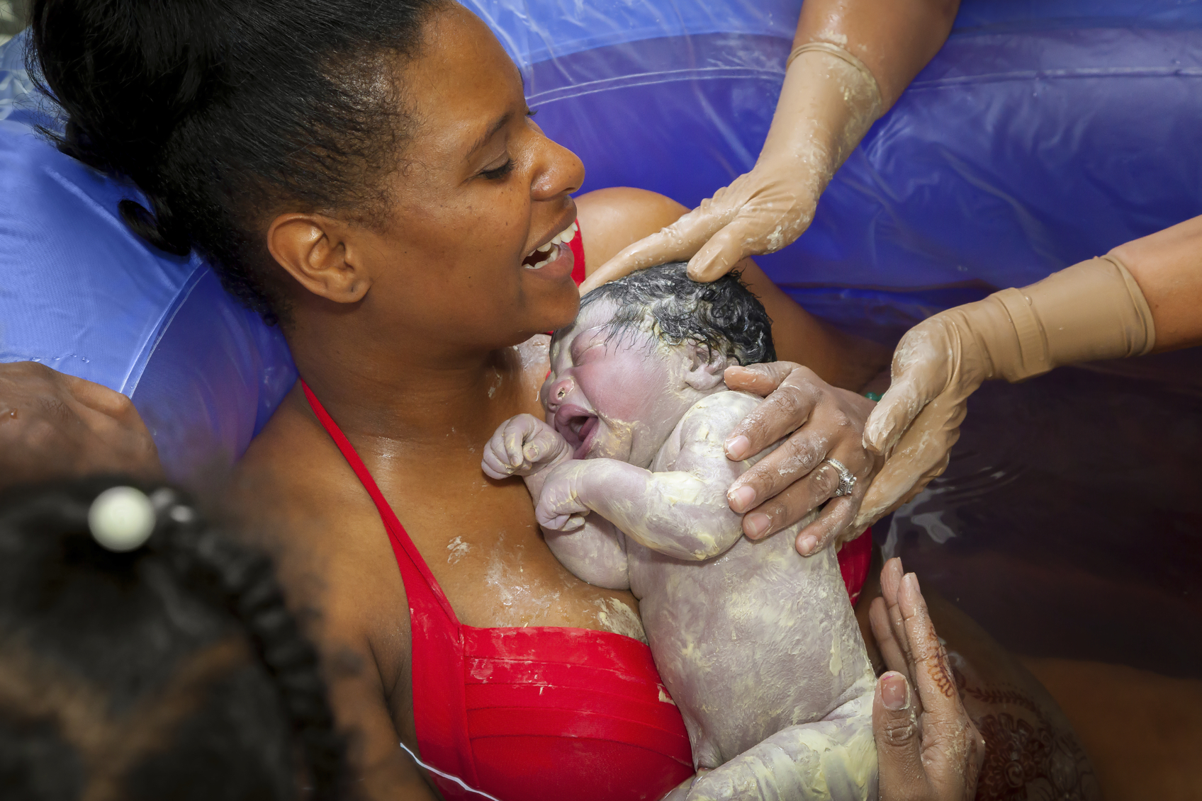 A happy African American mother holding her brand new baby girl after delivering her in a birthing pool at home. The baby is covered in vernix and the mom is laughing.