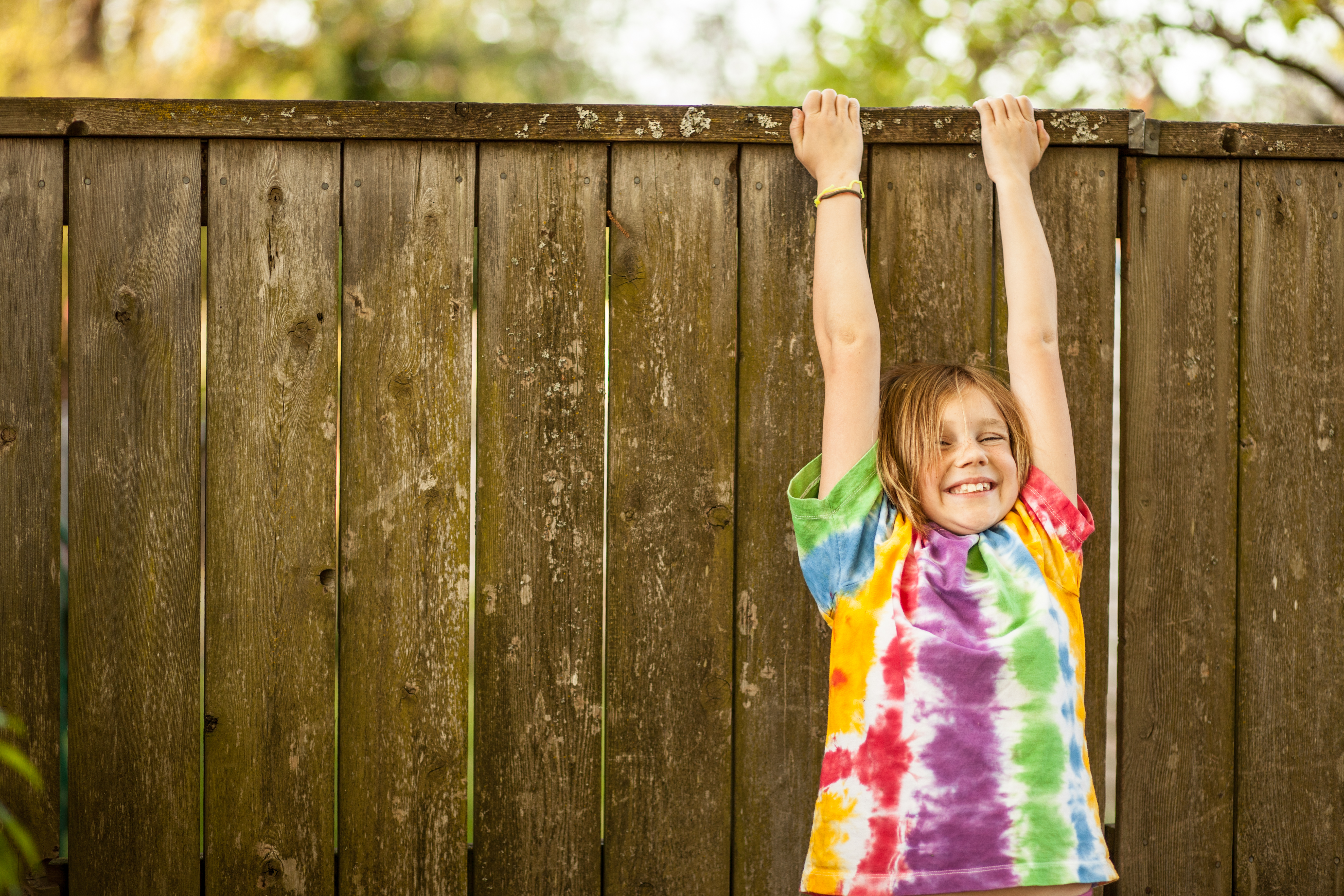 This is a color image of a young girl in a tie dyed T shirt she is smiling and hanging on a fence in her backyard.