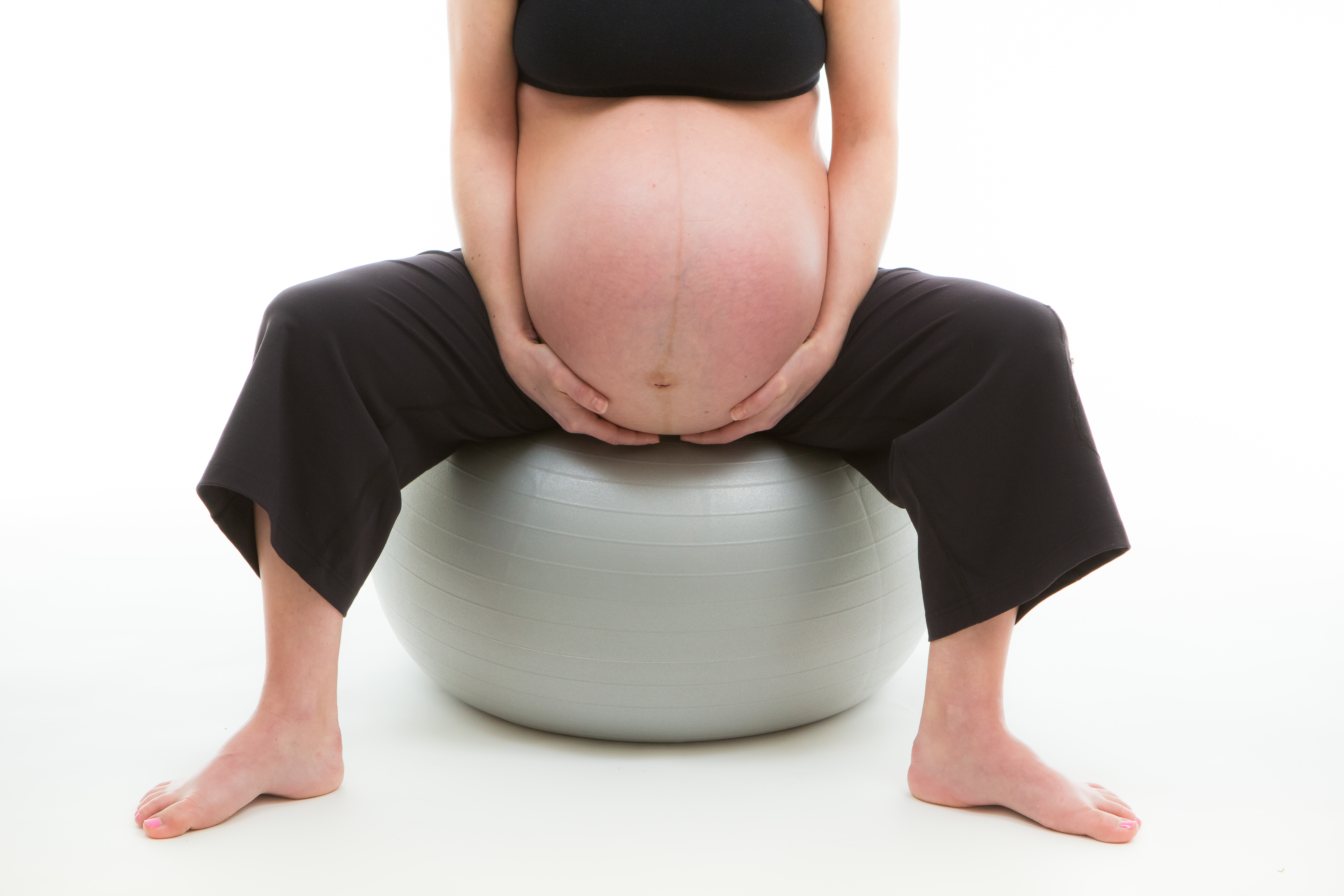 Pregnant woman in black pants and black sports bra on birthing ball on white background.