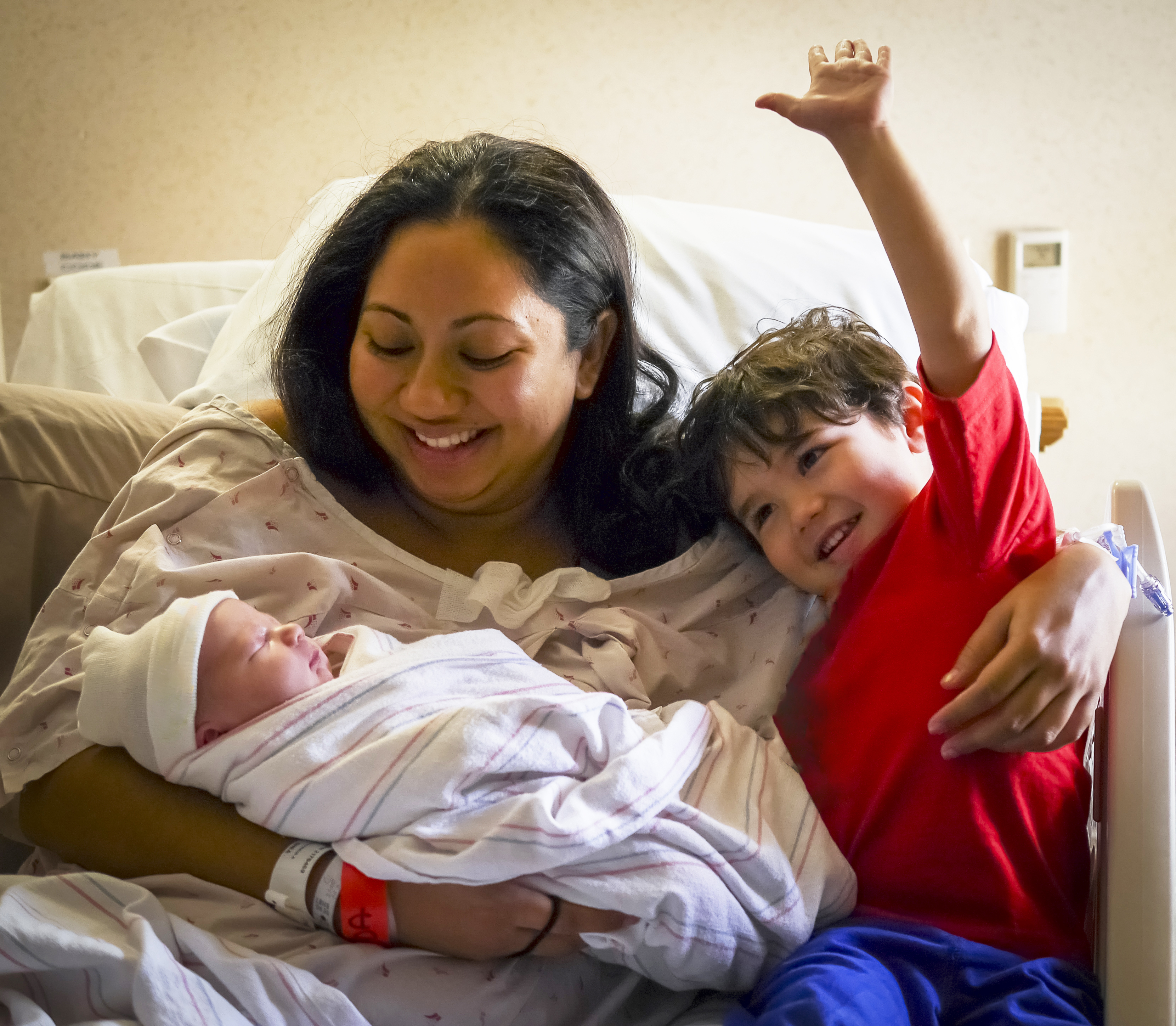 Mother in hospital bed with three year old son meeting his hours old brother for the very first time. These are the photographer's daughter-in-law and two grandsons. I removed the spiderman design from the 3 year old's shirt and the words on the baby's stocking cap.
